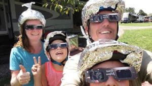 2 adults and 2 kids sitting together on chairs outside their RV with aluminum foil hats on their heads looking up at the blue sky