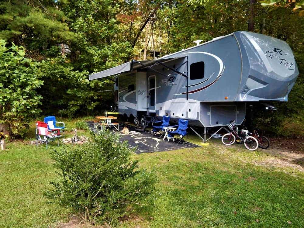open range 5th wheel in wooded setting with camp fire and chairs