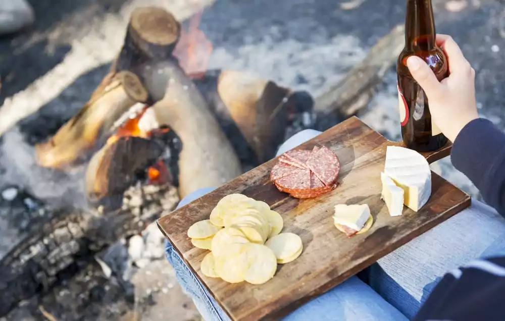camping food on a wooden board, with cheese, salami and crackers with a beer in front of the fire